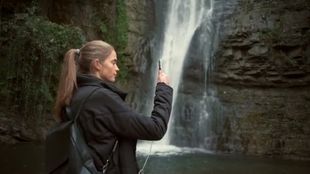 Mujer joven filmando un video de una cascada con un smartphone en cámara lenta — Vídeo de stock