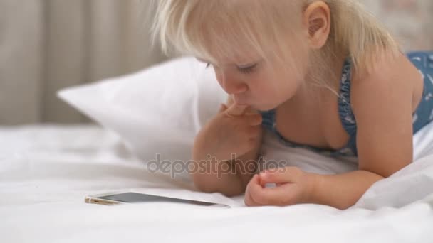 Dívka leží na bílé posteli a hraje smartphone s prstem v ústech — Stock video