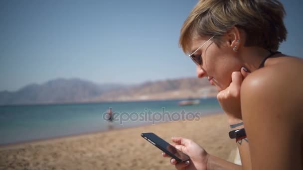 Красивая женщина сидит у моря и держит телефон замедленной съемки — стоковое видео