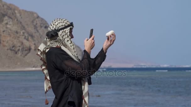 Hombre en una bufanda de pie en la orilla y haciendo una foto de una concha en cámara lenta — Vídeos de Stock