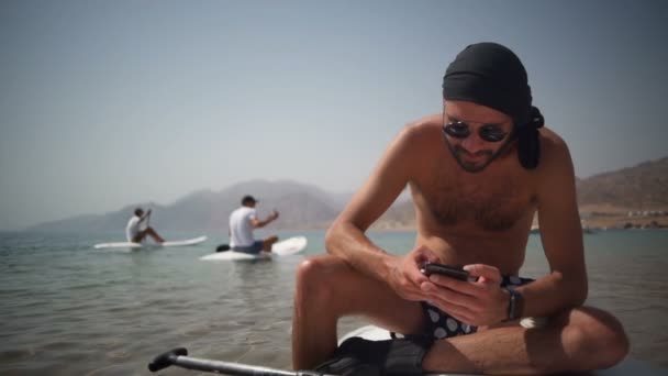 Человек в очках сидит на доске для серфинга и использует свой телефон замедленной съемки — стоковое видео