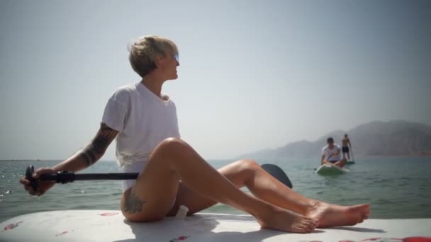 Unga blonda kvinnan sitter på paddling styrelsen och dans slowmotion — Stockvideo