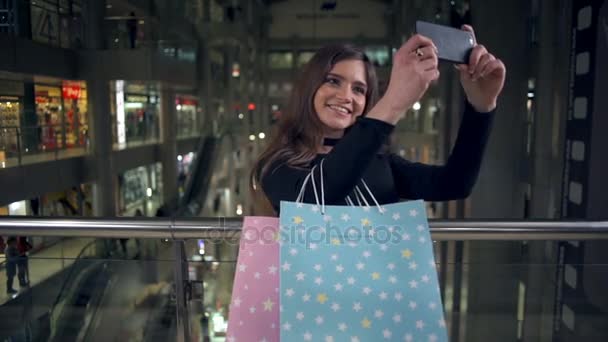 Jovem morena atraente com pacotes fazendo uma selfie no shopping câmera lenta — Vídeo de Stock