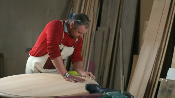 Carpenter leans on a table and polishes it with an abrasive block in workshop — Stock Video