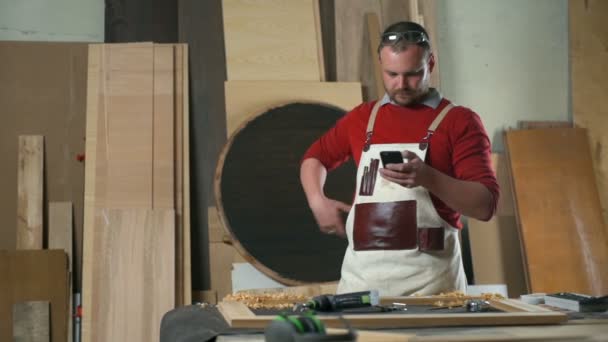 Carpinteiro vestindo um avental de madeira usando seu smartphone em uma oficina — Vídeo de Stock