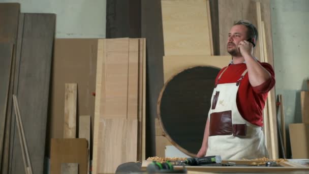 Carpintero con delantal de carpintería hablando por teléfono en un taller — Vídeo de stock