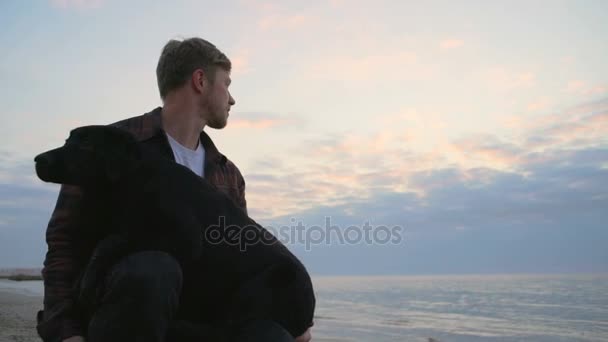 Молодий чоловік обіймає свого собаку біля моря і дивиться далеко повільно — стокове відео