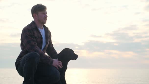 Jonge man zit met zijn hond aan zee en kijkt ver Slowmotion — Stockvideo