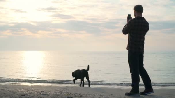 Молодой мужчина фотографирует свою любимую собаку у моря в замедленной съемке — стоковое видео