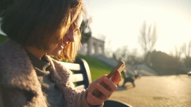 Mladá žena sedí na lavičce a pomocí její smartphone v parku closeup — Stock video