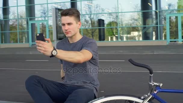 Young man sitting on the border and making a selfshot in front of a building — Stock Video