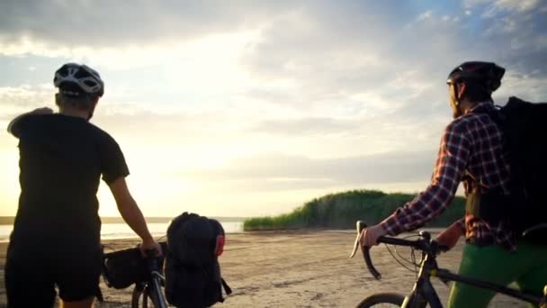 Dois ciclistas caminham até o mar com bicicletas à beira-mar mostram amanhecer câmera lenta rápida — Vídeo de Stock
