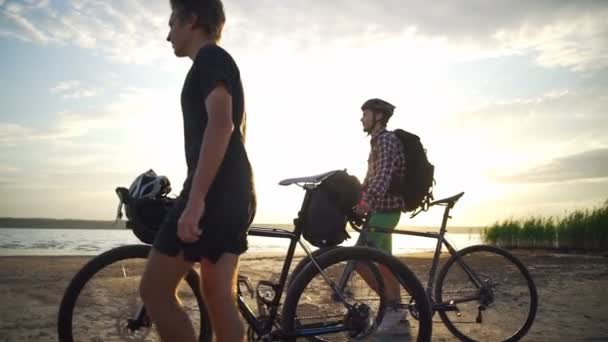 Dos jóvenes ciclistas caminan cerca del mar con bicicletas a orillas del mar amanecer cámara lenta rápido — Vídeo de stock