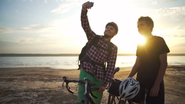 Dos jóvenes ciclistas hacen selfie en smartphone playa amanecer cámara lenta rápida — Vídeo de stock