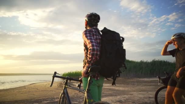 Dois jovens ciclistas apreciam o pôr do sol com bicicletas à beira-mar câmera lenta rápida — Vídeo de Stock