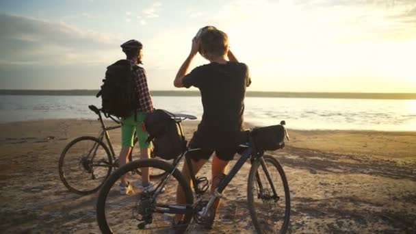 Два красивых велосипедиста наслаждаются закатом с велосипедами на берегу моря медленное движение быстро — стоковое видео
