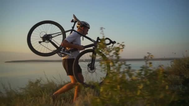 Fuerte ciclista lleva bicicleta en la costa del hombro vista al mar puesta de sol rápida cámara lenta — Vídeos de Stock