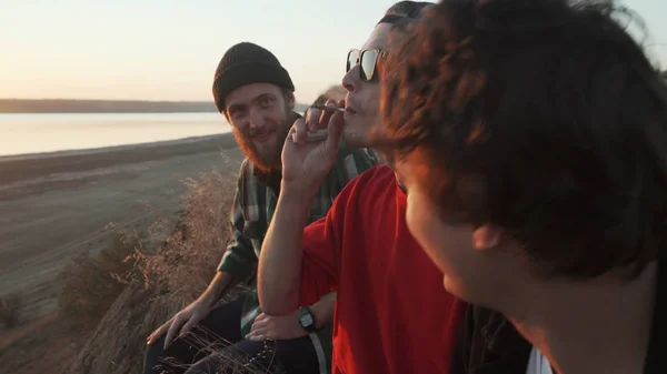 Друзья скейтбордисты курят травку на берегу моря на закате — стоковое фото