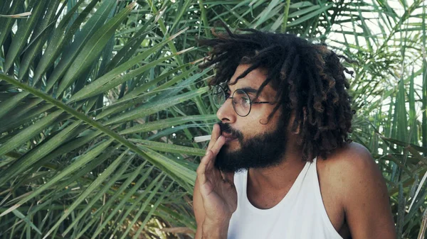 Homem preto com dreadlocks fumar maconha no fundo de folhas de palma — Fotografia de Stock