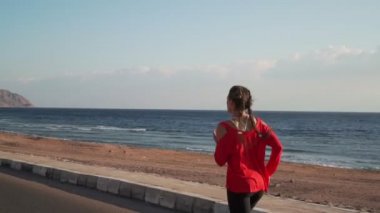 Athletic girl in a red sweater runs on the seashore at sunset slow motion