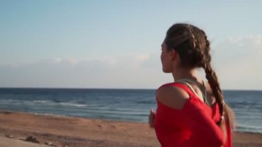 Athletic woman in a red sweater jogging by the ocean at sunrise slow motion