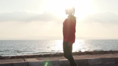 Young motivated girl is preparing to run on promenade at sunset near ocean