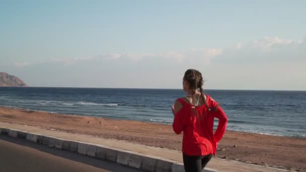 Chica atlética en un suéter rojo corre en la orilla del mar al atardecer cámara lenta — Vídeo de stock