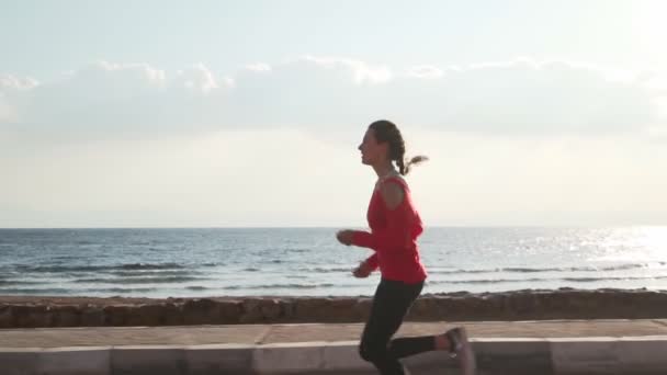 Ajustar morena mujer en un suéter rojo trotando por el océano al amanecer cámara lenta — Vídeo de stock