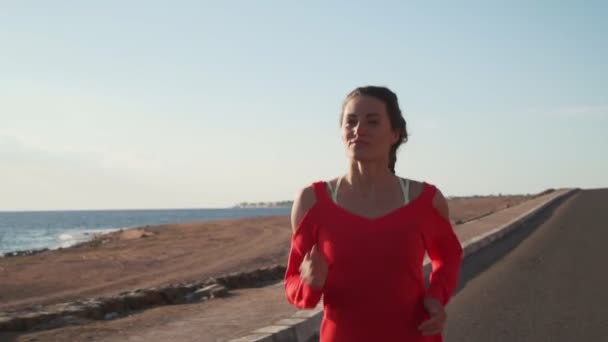 Joven mujer en forma corriendo en el camino vacío a la orilla del mar al atardecer cámara lenta — Vídeos de Stock