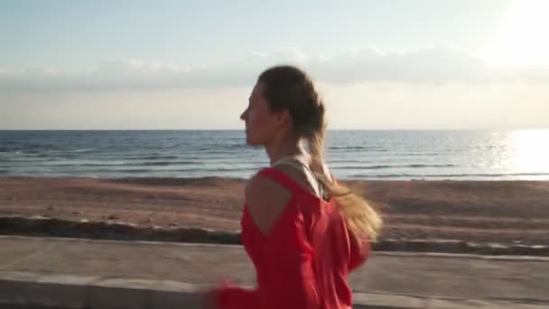 Beautiful young girl running on promenade at sunset near ocean — Stockvideo