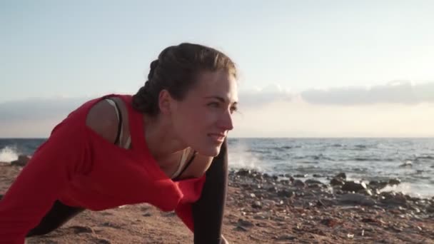Deniz kıyısında rüzgarlı bir sabahta çalışmadan önce esneme hareketleri yapan bir kadın. — Stok video