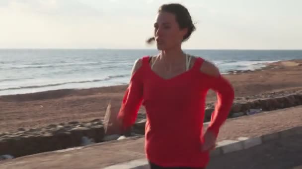 Athletic woman in sportswear running on promenade at sunset near ocean — Stockvideo