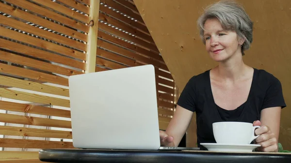 Laptop kullanan ve kafede kahve içen güzel, beyaz saçlı kadın.