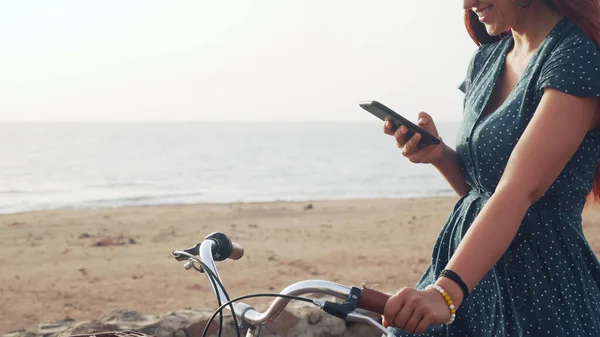 Bei capelli rossi femmina utilizzando il suo smartphone standig con la bicicletta in mare — Foto Stock