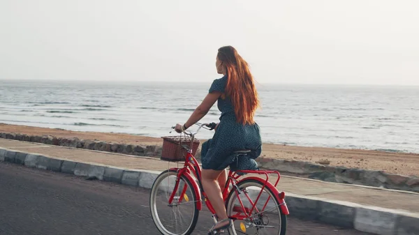 Yeşil elbiseli, güzel kızıl saçlı kadın sahil şeridi boyunca klasik bisiklet sürüyor.