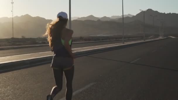 Vista trasera de la joven chica delgada trotando en la carretera en las montañas al atardecer — Vídeo de stock