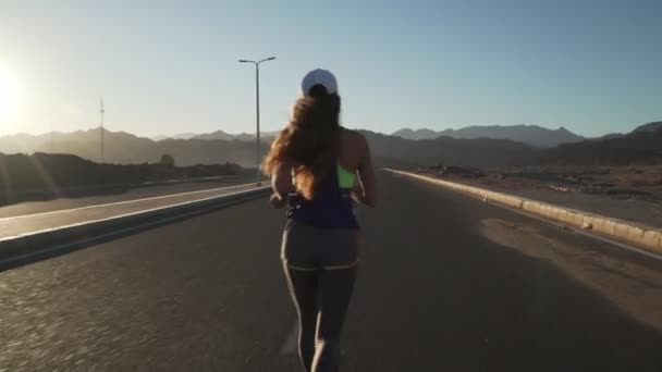 Vista posterior de la joven chica motivada trotando en la carretera al atardecer — Vídeos de Stock