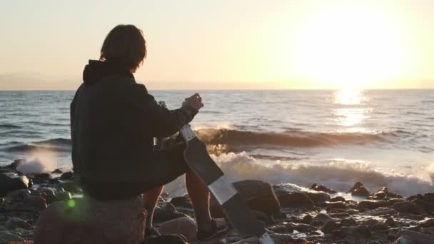 Мальчик со скейтбордом пьет горячий чай из термоса на берегу моря на закате — стоковое видео