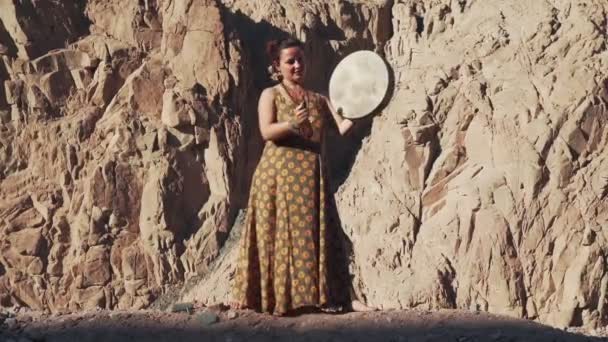 Young caucasian woman in colorful dress playing tambourine with mountains at background — Stock Video