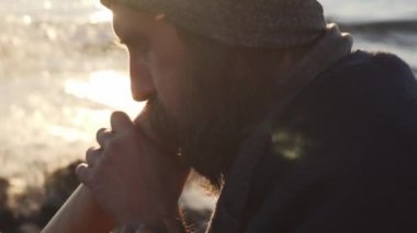Yakın plan yakışıklı adam deniz kıyısında gün ışığında didgeridoo çalıyor.