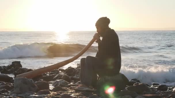 Veduta dell'uomo seduto sulla riva del mare alla luce del sole e suonare lo strumento didjireedoo — Video Stock