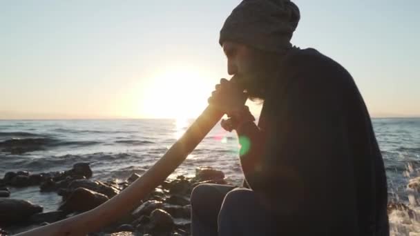 Barbudo tocando su didgeridoo instrumento australiano bajo el sol en la orilla del mar — Vídeo de stock