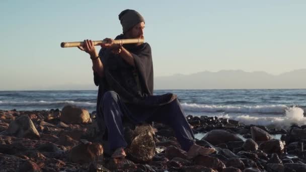 Bonito homem barbudo tocando instrumento indiano Bansuri ao sol na praia — Vídeo de Stock