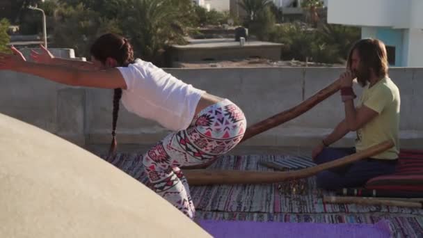 Girl practice yoga asana while man plays didgeridoo instrument at sunset rooftop — Stock Video