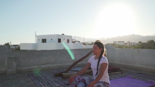 Молода дівчина практикує медитацію, а чоловік грає діджериду в сонячних відблисках на даху — стокове відео