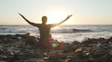 Bir kadının namaste pozunda oturup gün batımında deniz kenarında meditasyon yapması.