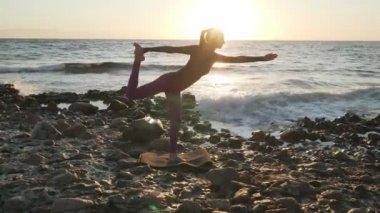 Genç bir dişi, deniz kıyısında yoga pozu vererek yay kaldırıyor.