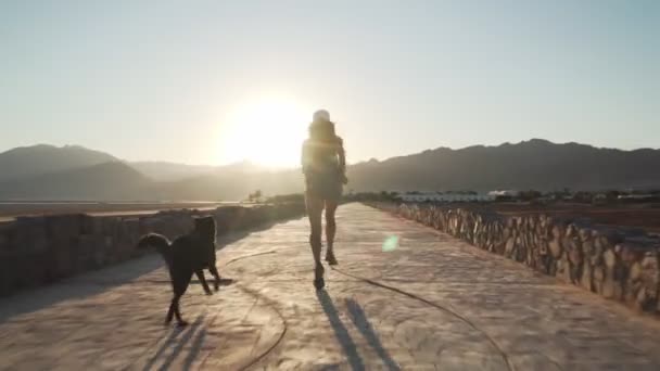 Vista posteriore femminile corre con il suo cane divertente in strada asfaltata deserto asciutto all'alba — Video Stock