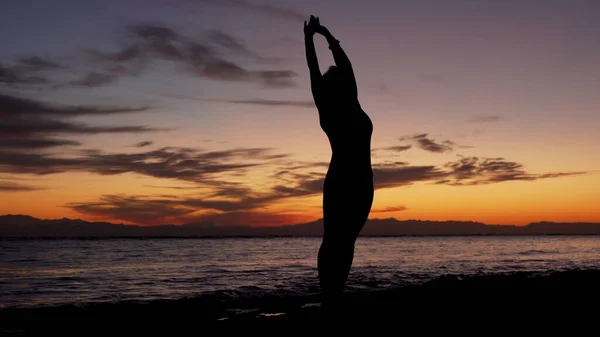 La silueta de la muchacha practica yoga en la orilla del mar en la puesta de sol colorida brillante —  Fotos de Stock