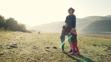 Young caucasian mother hugging with her blonde daughters near lake and mountains